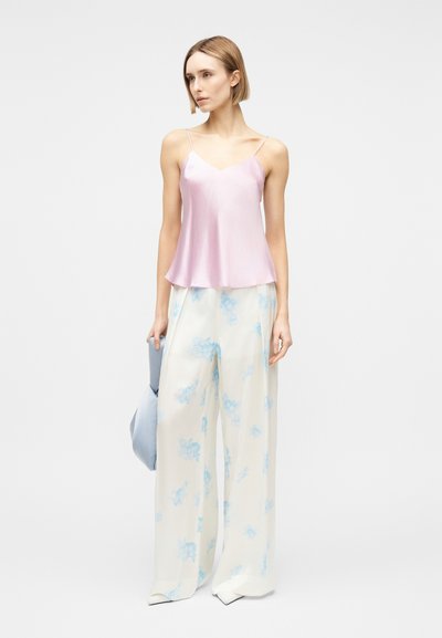 Γυναίκα που φοράει ανοιχτό ροζ σατέν καμισόλ και λευκό παντελόνι με φαρδιά μπατζάκια και μπλε λουλουδένια σχέδια, κρατώντας ένα διπλωμένο μπλε σακάκι στο αριστερό της χέρι.