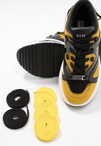 Basket jaune et noire avec une plaque en métal « Steve Madden », présentée avec deux paires de lacets noirs et quatre paires de lacets jaunes à côté.