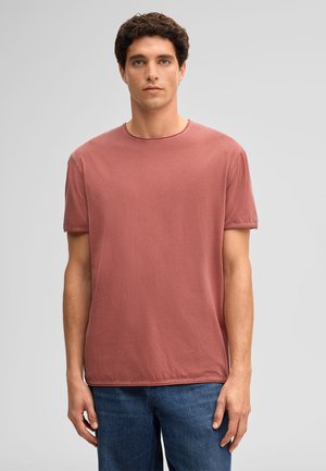 Jeune homme aux cheveux courts et bouclés portant un T-shirt à manches courtes rose fané et un jean bleu, debout devant un fond gris uni.