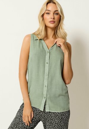 Femme blonde portant un chemisier vert clair sans manches avec des boutons et un pantalon noir à motif abstrait blanc, posant devant un fond uni.