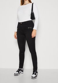 Vaqueros pitillo negros con bolsillos delanteros con cremallera y textura acanalada en los muslos, combinados con una camiseta blanca de manga larga y zapatillas negras.