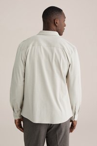 Šviesiai smėlio spalvos ilgomis rankovėmis marškiniai iš tekstūruotos medžiagos, su sagomis užsegamais rankogaliais ir smailiomis apykaklėmis, parodyti iš nugaros neutralaus fono fone.