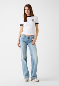 Biała koszulka z krótkim rękawem z czarnym wykończeniem i logo "G", zestawiona z jasnoniebieskimi dzwonami i srebrnymi sneakersami. Gładka tkanina i casualowy design.