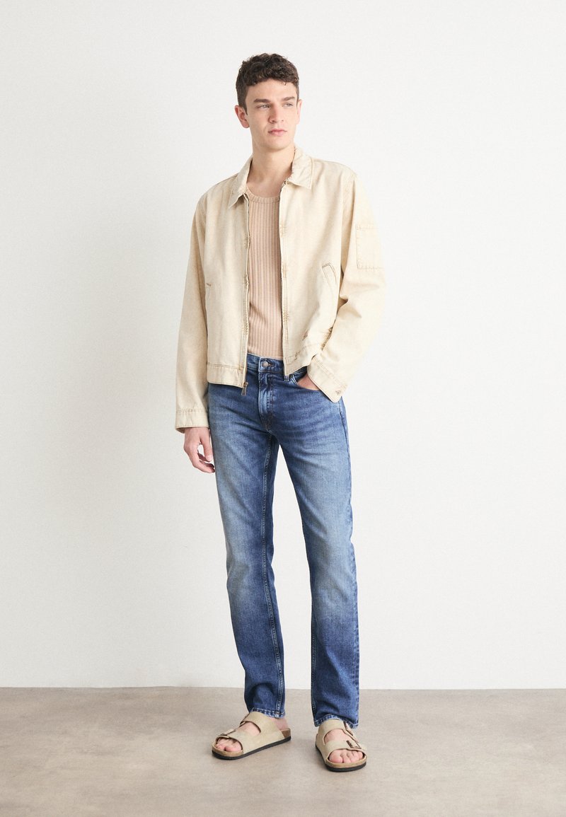 Beige Denimjacke, gerippter beiger Pullover, blaue Jeans und helle Sandalen in Tan. Glatte Textur, lässige Passform und schlichtes Design.