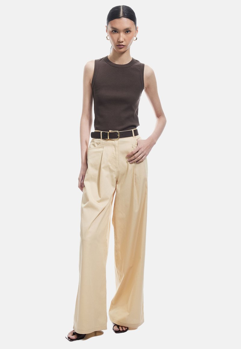 Bruine mouwloze geribbelde top gecombineerd met lichte, beige wijde broek. Zwarte riem met gouden gespaccenten. Model staat tegen een witte achtergrond.