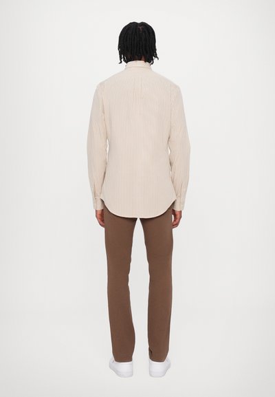 Juostuotas smėlio spalvos marškiniai su ilgomis rankovėmis, išlenktu kraštu, derinami su ruda prigludusia kelnėmis ir baltomis sportinėmis bateliais.