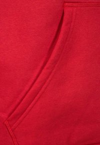 Haut à capuche rouge avec une texture douce, présentant une poche avant et des accents cousus. Le matériau semble durable et léger.