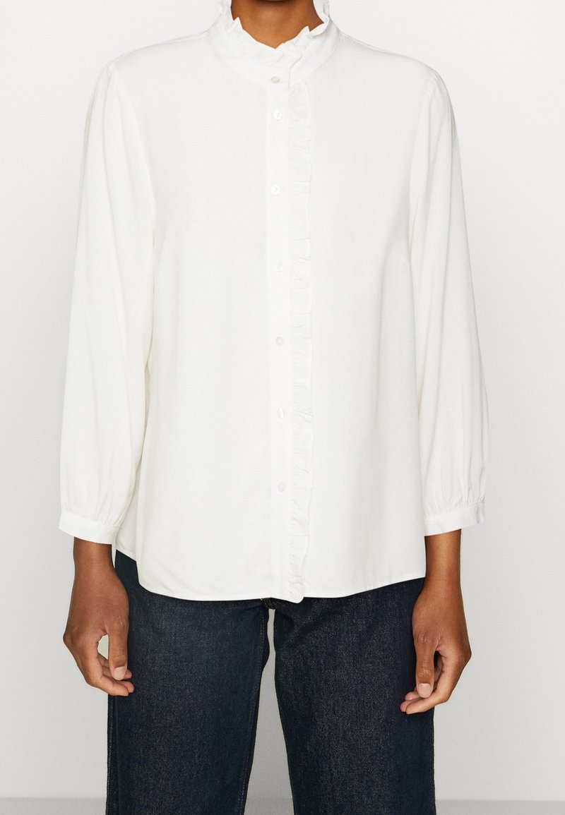 Blouse blanche à manches longues avec patte de boutonnage à volants et col, portée avec un jean bleu foncé, vue du cou jusqu'aux hanches.