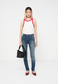 Mujer joven vestida con una camiseta sin mangas blanca con ribete rojo, jeans azules y zapatos planos rojos, sosteniendo un bolso negro frente a un fondo blanco.