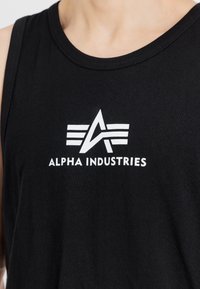 Μαύρη αμάνικη μπλούζα φτιαγμένη από βαμβάκι, με λευκό λογότυπο "Alpha Industries" και γεωμετρικό σχέδιο στο μπροστινό μέρος.