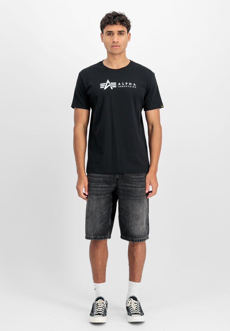 Zwarte katoenen T-shirt met wit "ALPHA INDUSTRIES" logo, gecombineerd met donkere denim shorts. Model draagt witte sokken en zwarte sneakers.