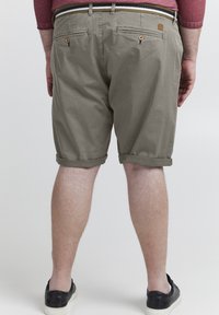 Shorts kaki avec un ourlet roulé, fabriqués en coton, dotés de deux poches arrière avec des fermetures à bouton, portés avec des baskets noires.