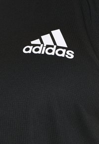 Logo blanc Adidas avec trois bandes diagonales sur fond de tissu noir texturé.