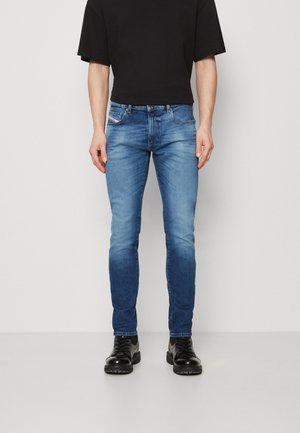 Homme portant un jean slim bleu, un t-shirt noir court et des bottes noires brillantes à lacets, debout devant un fond clair uni.