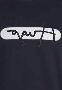 Sudadera de color navy con un parche ovalado blanco y texto bordado en negro. El material tiene una textura suave, con detalles de cuello de canalé.