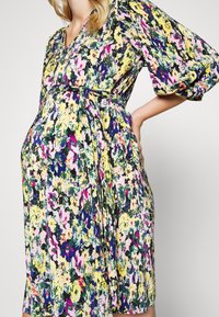Femme enceinte portant une robe fleurie colorée avec des manches bouffantes et une ceinture nouée à la taille, debout avec une main sur la hanche.