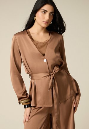 Donna con lunghi capelli scuri che indossa una giacca avvolgente in raso marrone e pantaloni abbinati, accessoriata con braccialetti spessi e orecchini d’oro.