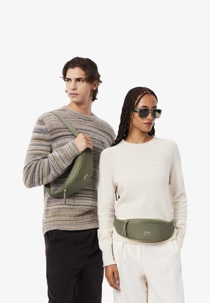 Marsupio verde di forma arrotondata con chiusura a zip, indossato da modelli. Un modello con un maglione a righe, l'altro con un maglione color crema.