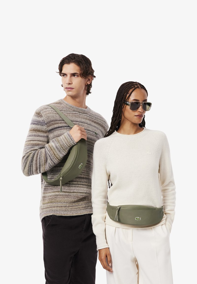 Marsupio verde di forma arrotondata con chiusura a zip, indossato da modelli. Un modello con un maglione a righe, l'altro con un maglione color crema.