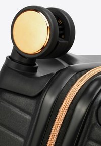 Czarny narożnik walizki na kółkach z teksturowaną powierzchnią, z złotym akcentem na kole i detalem zamka w kolorze różowego złota.