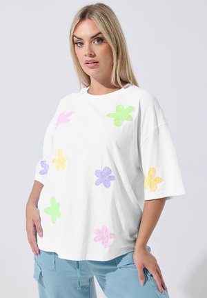 Blondynka w luźnej białej koszulce z kolorowymi pastelowymi kwiatowymi wzorami i jasnoniebieskich spodniach, stojąca na jednolitym tle.