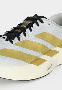 Chaussure de sport blanche avec des bandes dorées, des lacets noirs et la technologie de semelle "Lightstrike Pro" visible sur le côté.