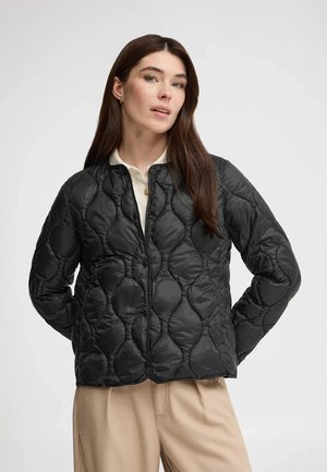 Kvinde med langt brunt hår iført en sort quiltet jakke over en hvid skjorte og beige bukser, stående med hænderne bag ryggen.