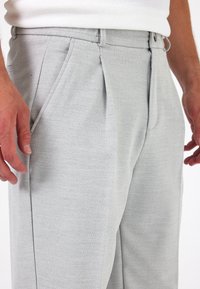 Lichtgrijze broek van een gestructureerde stof, met geplooide voorkant, zijzakken en riemlussen. Close-up van de taille en heupen.
