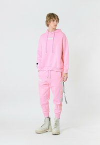 Sudadera con capucha rosa y bolsillo frontal, joggers rosa a juego con parches laterales, combinados con botas de combate blancas. Textura de tela suave.