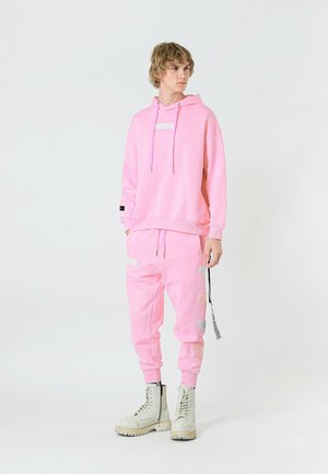 Sudadera con capucha rosa y bolsillo frontal, joggers rosa a juego con parches laterales, combinados con botas de combate blancas. Textura de tela suave.