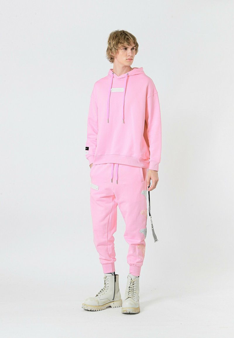 Sudadera con capucha rosa y bolsillo frontal, joggers rosa a juego con parches laterales, combinados con botas de combate blancas. Textura de tela suave.