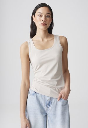 Young woman with long dark hair wearing a beige tank top and light blue jeans, standing with one hand in pocket against a plain background.