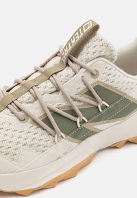 Primer plano de unas zapatillas beige y verdes con parte superior de malla, cordones beige, tirador con marca y suela de goma texturizada.