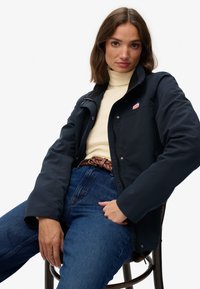 Marineblauwe softshelljas met drukknoopjes, twee zakken en een logo patch. Draagt over een crèmekleurige coltrui en gecombineerd met blauwe jeans.