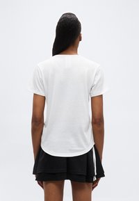 Άτομο με μακριά σκούρα μ αλλιά που φοράει απλό λευκό κοντομάνικο πουκάμισο και μαύρη πολυεπίπεδη φούστα, απεικονιζόμενο από πίσω μπροστά σε απλό φόντο.