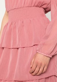 Vestido rosa em camadas com uma cintura franzida, mangas longas e um tecido leve e texturizado. O corpete é ligeiramente pregue.