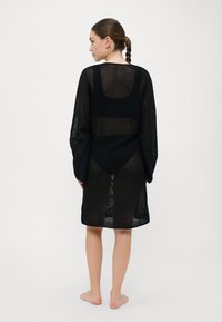 Black, sheer knit cover-up dress with long sleeves, featuring a straight cut and side slits, worn over black swimwear.