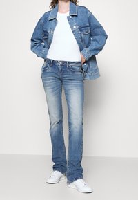 Veste en denim bleu clair à coupe oversize, associée à un jean droit bleu clair et des baskets blanches ; texture lisse, design classique à cinq poches.