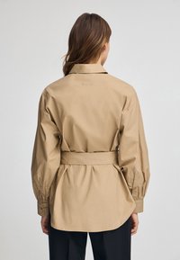 Helle braune Hemdjacke mit Gürtel, strukturiertem Kragen, langen Puffärmeln und geschwungenem Saum. Der Stoff wirkt glatt und leicht glänzend.