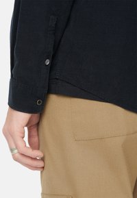 Camicia in cotone a coste blu scuro con tessuto testurizzato, polsini con bottone e orlo angolato, indossata sopra pantaloni khaki con una tasca posteriore visibile.