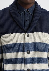Strickjacke mit navy, weißen und blauen Streifen, rippigem Muster, Schalkragen und schwarzen Knöpfen vorne.