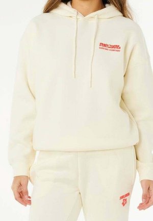 Femme portant un sweat à capuche couleur crème et un pantalon assorti avec des logos rouges "Rip Curl Surfing Company" sur la poitrine et la cuisse.