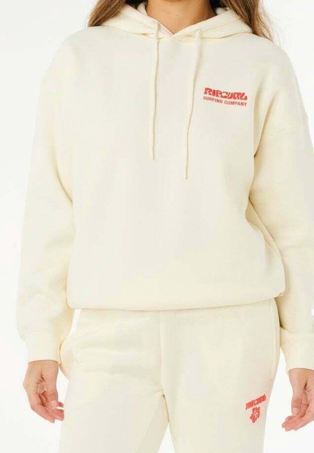 Femme portant un sweat à capuche couleur crème et un pantalon assorti avec des logos rouges "Rip Curl Surfing Company" sur la poitrine et la cuisse.