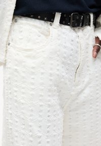 Pantaloni di jeans bianchi con un motivo testurizzato a zigzag, dotati di bottoni metallici e una cintura nera con borchie per un tocco in più.