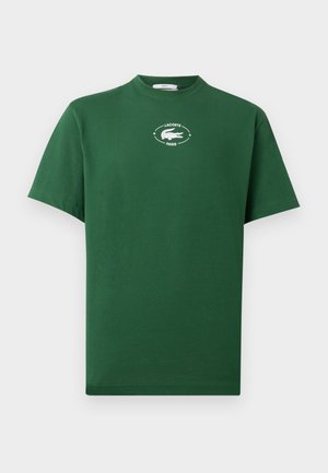 Homme et femme portant des t-shirts verts Lacoste et des pantalons de survêtement blancs, debout contre un mur blanc, tous deux face à l'avant.