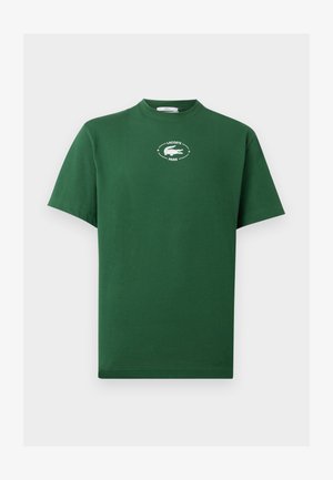 Homme et femme portant des t-shirts verts Lacoste et des pantalons de survêtement blancs, debout contre un mur blanc, tous deux face à l'avant.