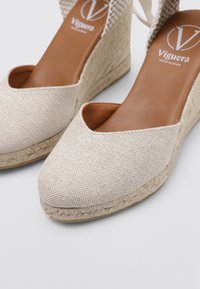 Sandalias de cuña de tela beige con un diseño de punta abierta, detalles de cuerda de yute y una suela lisa color tostado. Presenta correas en los tobillos para un ajuste seguro.