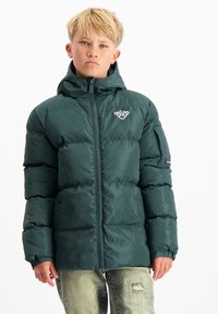 Groene gewatteerde jas met een capuchon, vooraan met een rits en elastische manchetten. Bevat een logo op de borst en een zak op de mouw. G gedragen met vervaagde jeans.