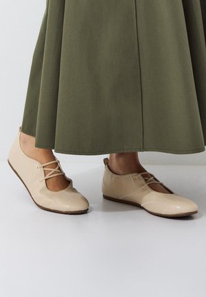 Pieds portant des chaussures plates beiges à lacets, debout sur un sol blanc avec une longue jupe vert olive visible.