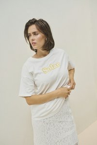 T-shirt en coton blanc avec "Solar" brodé en jaune. Associé à une jupe en dentelle blanche, ornée de motifs floraux et avec une silhouette ajustée.